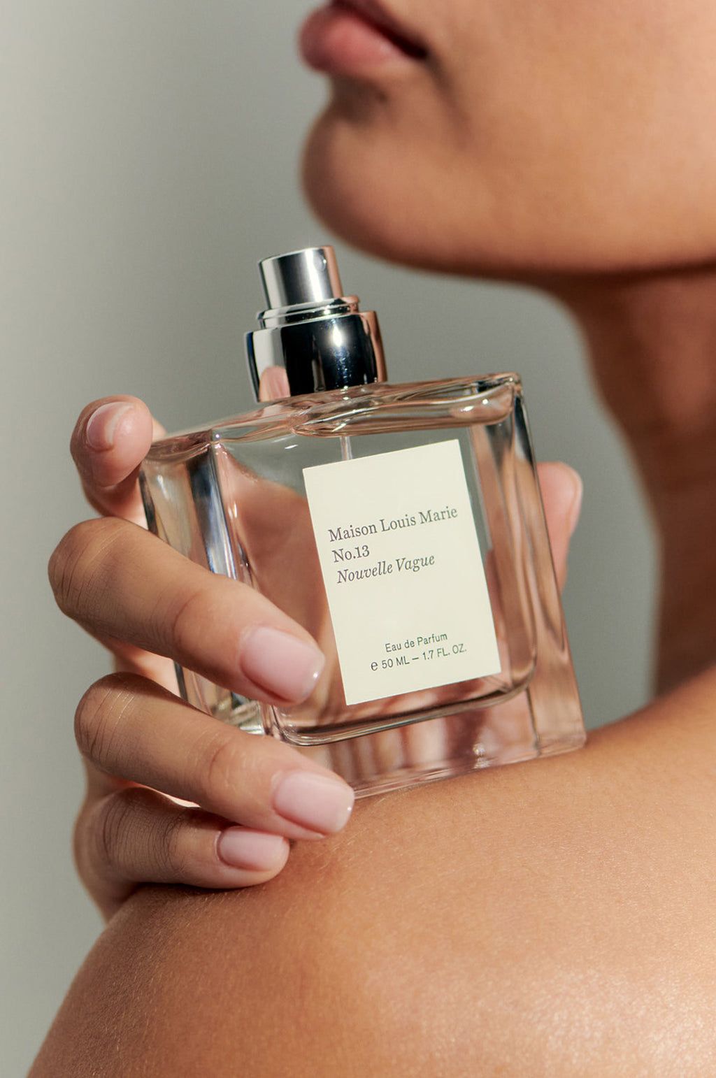 Close-up of person holding Maison Louis Marie No.13 Nouvelle Vague Eau de Parfum bottle near shoulder.